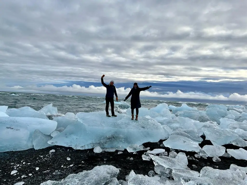 Two people with arms raised on ice blocks on a black sand beach