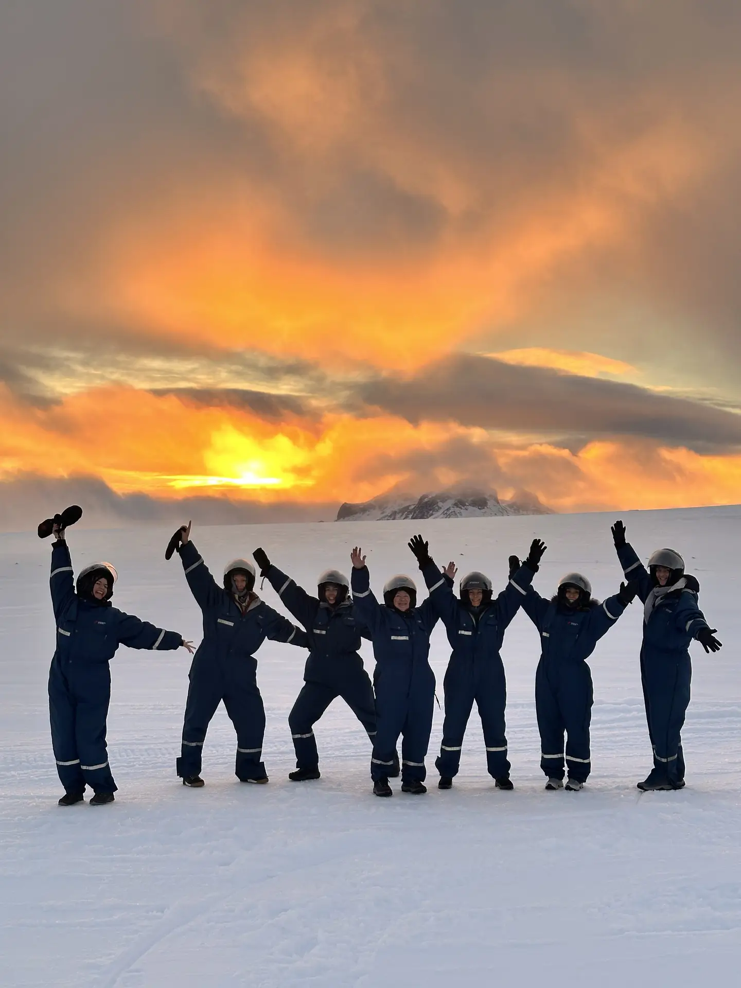 Group in snowsuits with arms raised under an orange sunset