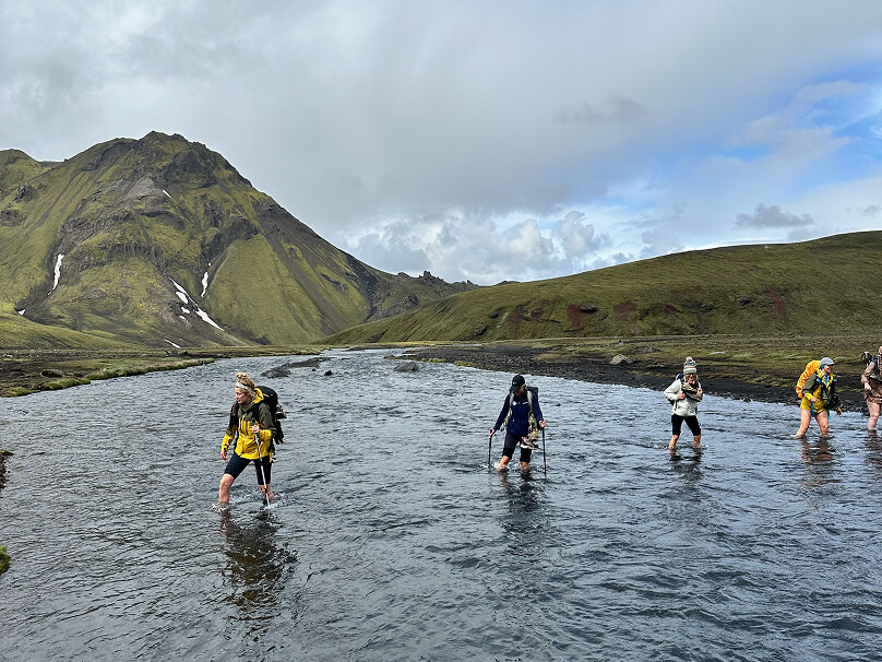 Hikers wading across a wide river in a green valley