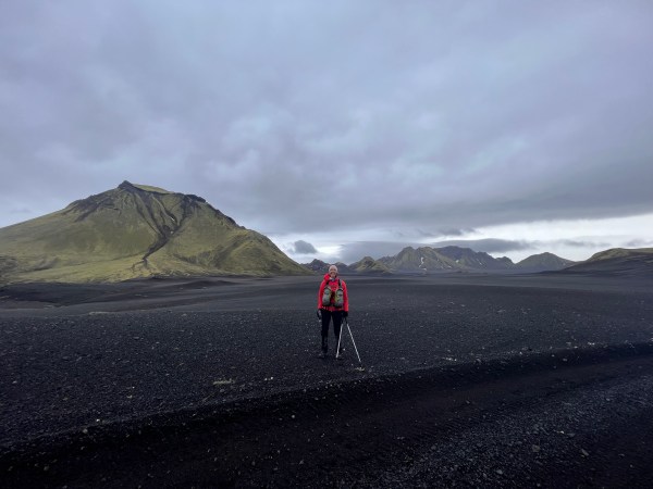 Hiker with poles on a black sand plain beneath green mountains